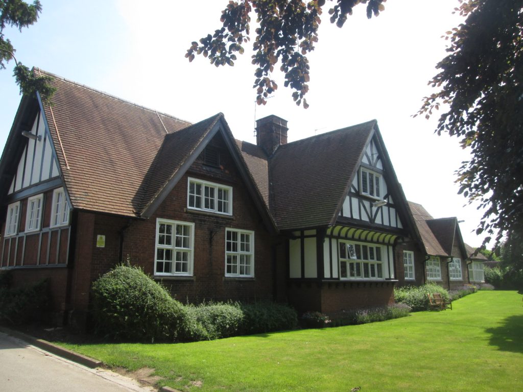 St Monica's Hospital; a large brick building with gabled roofs, white-framed windows, and black-and-white timber detailing, set on a lawn with shrubs and trees.